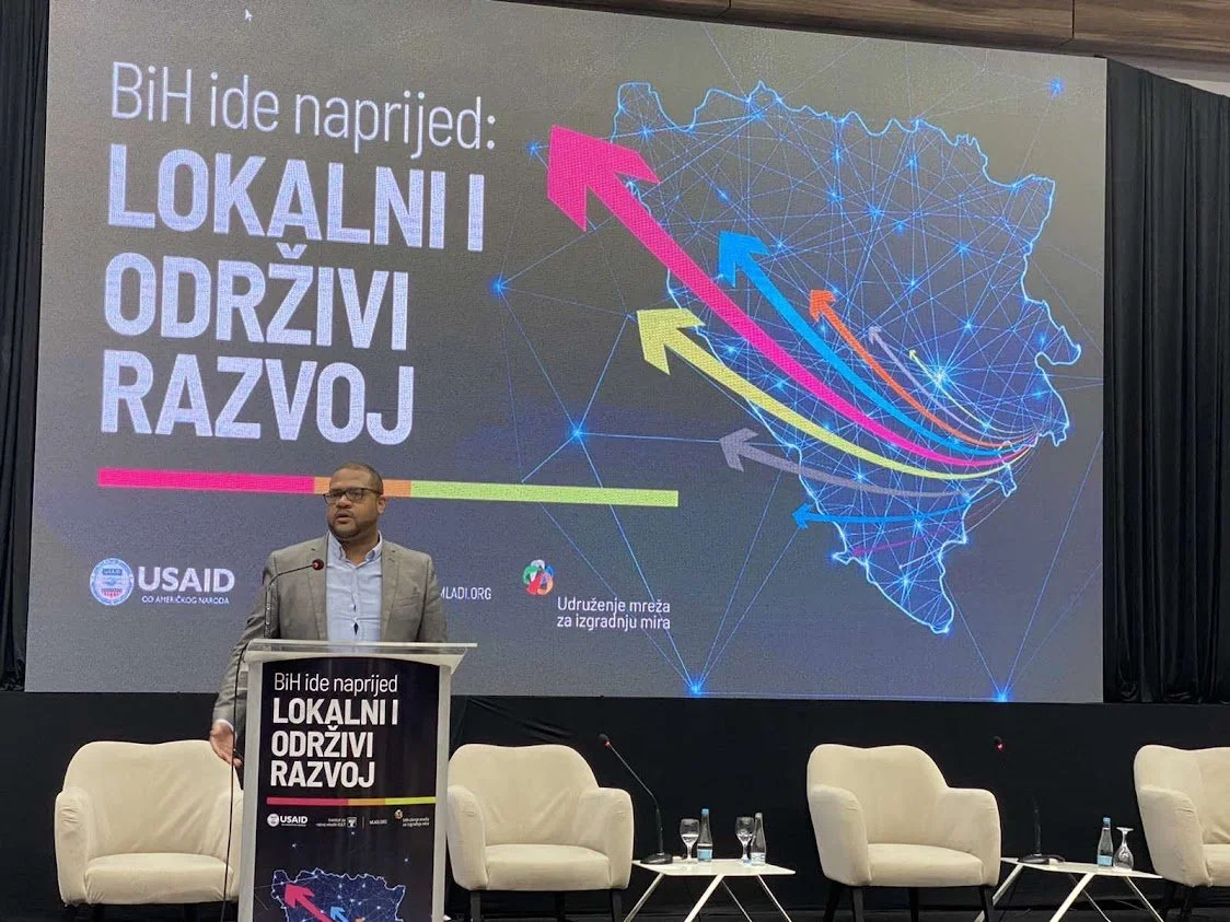 A man standing at a podium during a presentation, with a large screen behind him displaying a map of Bosnia and Herzegovina and colorful arrows, at an event focused on local sustainable development in Bosnia and Herzegovina.
