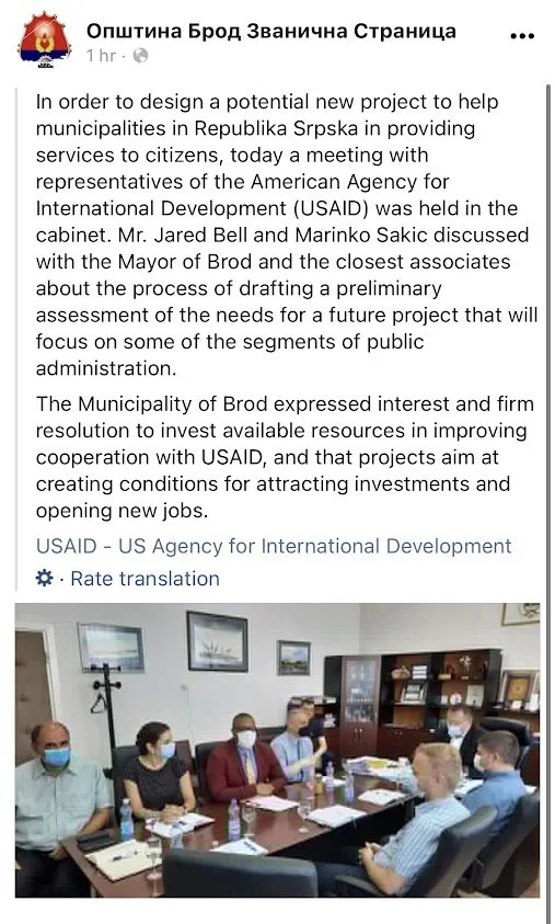 Meeting room with seven people wearing masks, sitting around a table, discussing documents and using laptops.