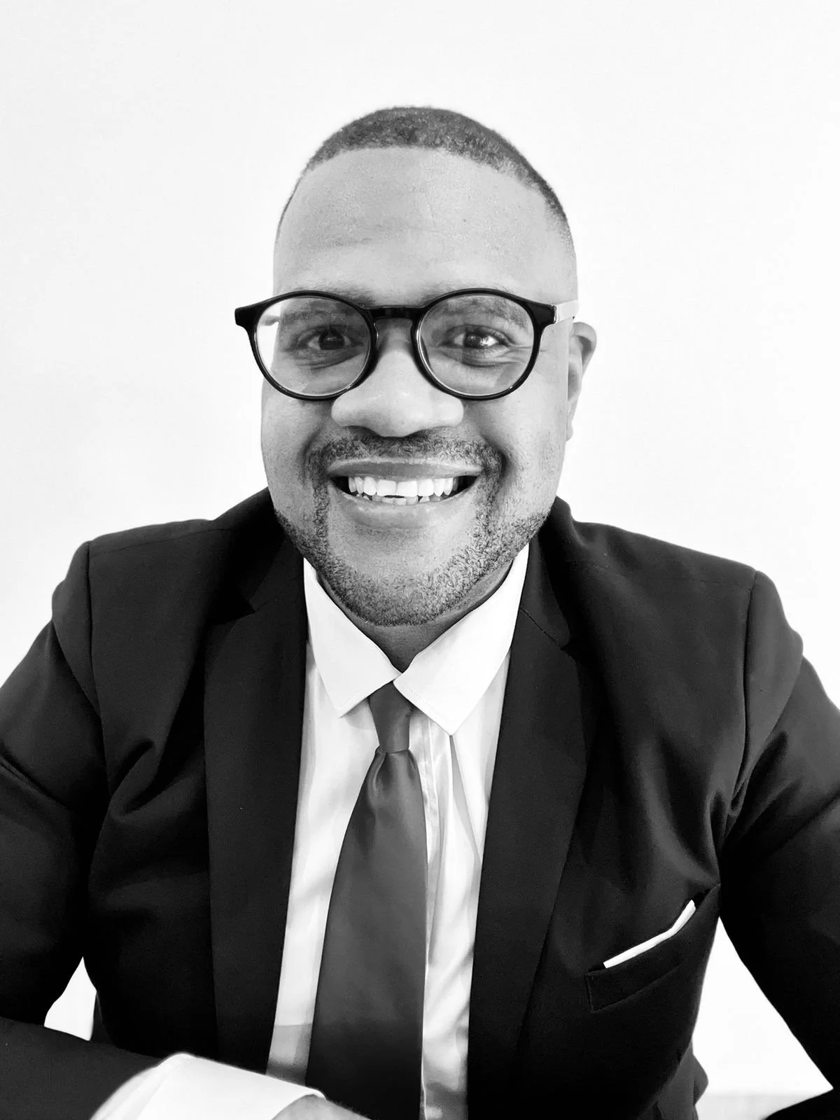 Black and white photo of a smiling man wearing glasses, a dark suit, white shirt, and tie, sitting against a plain white background.