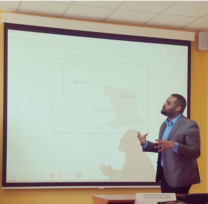 Man in a suit giving a presentation about the Swazi people, with a map showing the regions of Siwa, Nizwa, and Palmyra projected on a screen.