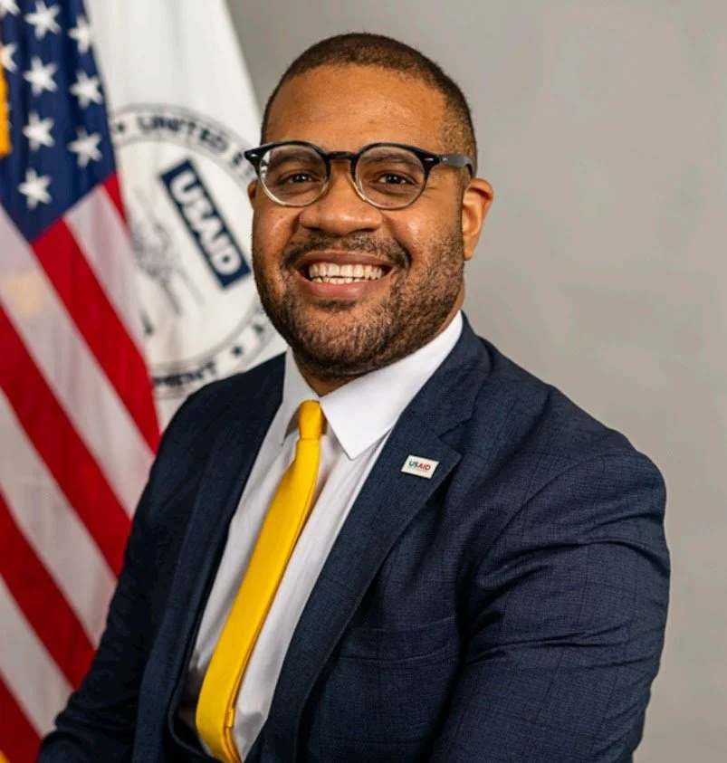 A smiling man in a suit and yellow tie, wearing glasses with a lapel pin, standing in front of an American flag and a U.S. Army flag.