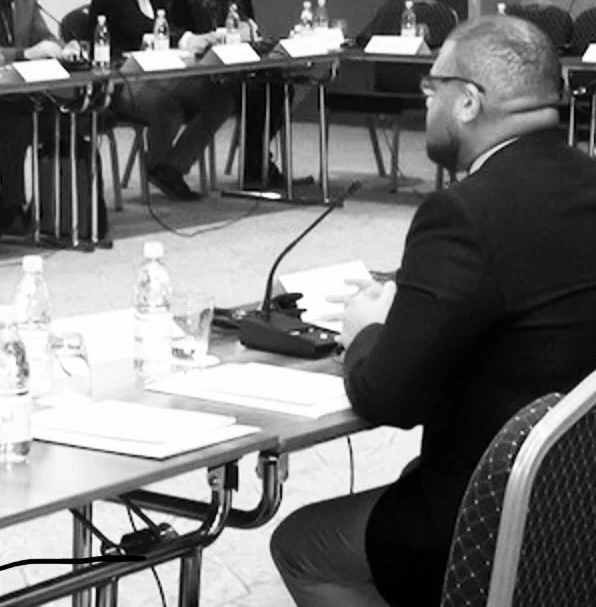 A man in a suit is seated at a conference table, holding a paper, with microphones and water bottles on the table, at a formal meeting or conference.