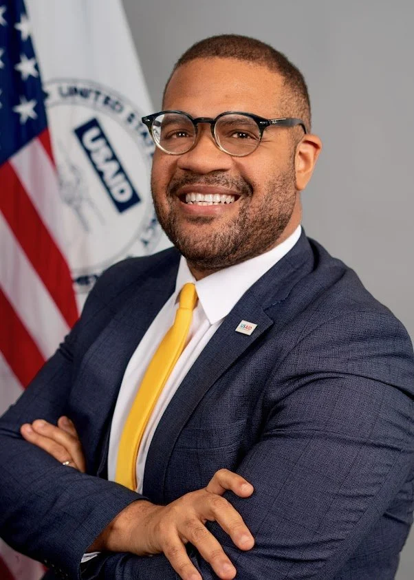 A smiling man in a suit with glasses, standing with crossed arms in front of a United States flag and a U.S. Department of Education flag.