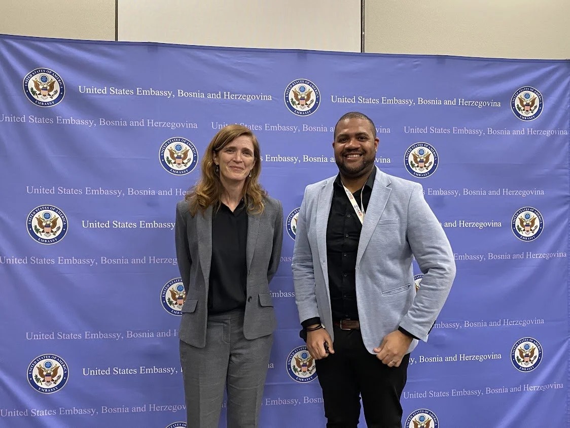 Two people, a woman and a man, standing in front of a blue backdrop with the U.S. Embassy seal and text, smiling for the photo.