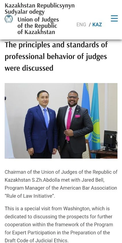 Two men in suits standing in front of a white wall with flags in the background, discussing principles and standards of professional behavior of judges.