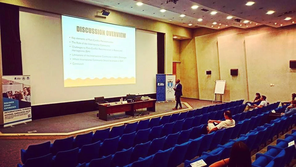 A speaker giving a presentation on a stage in a conference room with a large screen, titled 'Discussion Overview', and a few attendees sitting in blue chairs.