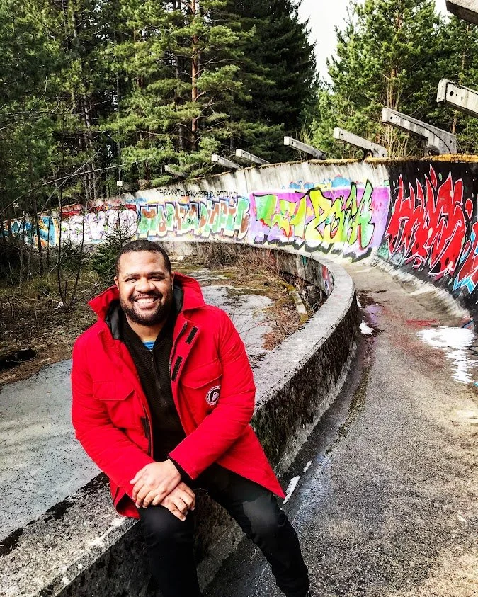 A smiling man in a red jacket sitting on a concrete ledge with graffiti, in an outdoor area surrounded by trees.
