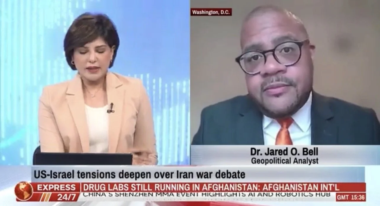 News broadcast with female anchor on the left and male guest Dr. Jared O. Bell on the right, discussing US-Israel tensions over Iran war debate. Female anchor has short brown hair, beige blazer, and white top. Male guest wears glasses, suit, white shirt, and orange tie, with a label indicating he is a geopolitical analyst in Washington, D.C.