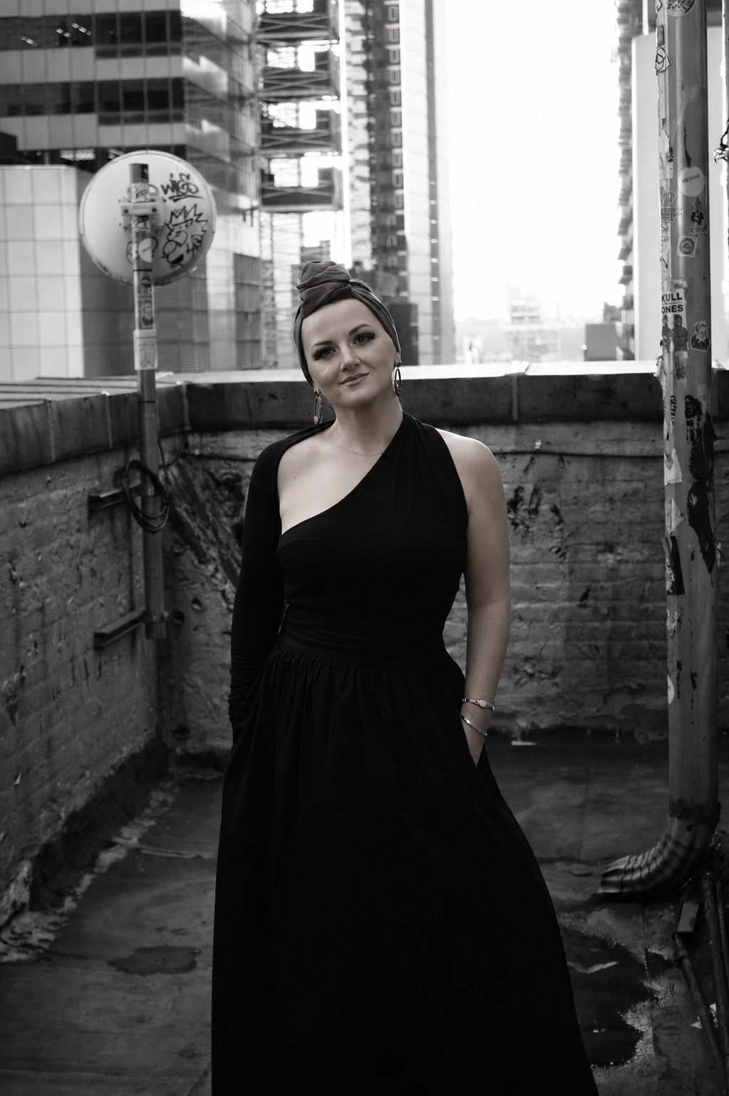 A woman standing in an urban rooftop setting, dressed in a black dress with a one-shoulder design, with her hands in her pockets and wearing large hoop earrings, with city buildings in the background.