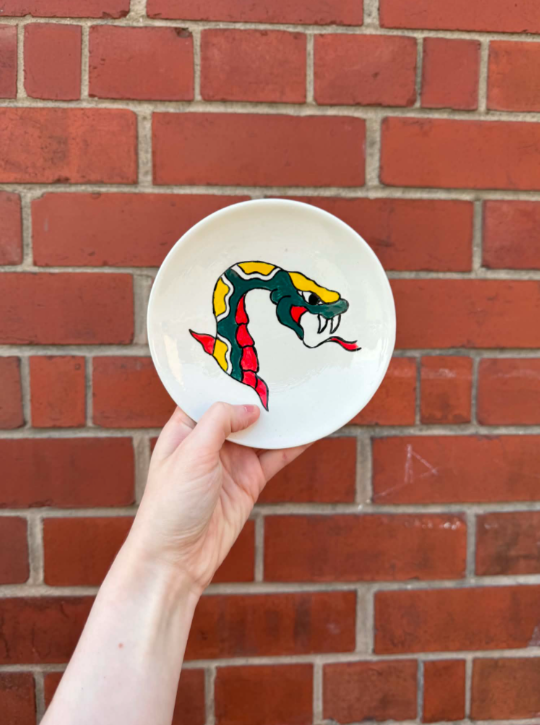 White ceramic plate with a colorful cartoon snake design, held by a hand against a red brick wall background.