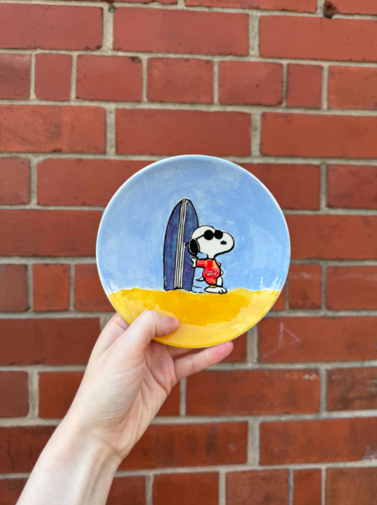 A circular painted ceramic plate featuring Snoopy with sunglasses and a red striped shirt standing on a sandy beach near a surfboard against a blue sky background. The plate is held in front of a brick wall.