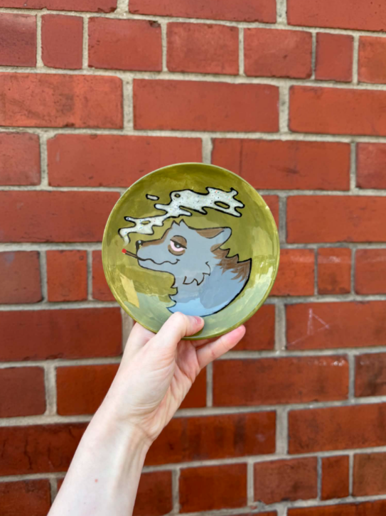 A hand holding a round green ashtray with a cartoonish dragon design against a red brick wall.