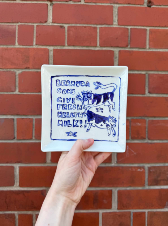 A person holding a square white plate with a hand-painted blue design of a cow and a goat, and text that reads "Bermuda Goes Gives Fresh Healthy Milk!" against a brick wall background.