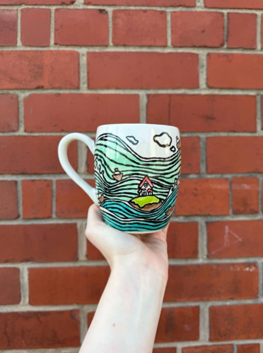 Hand holding a coffee mug with a colorful landscape design of a house on a hill, trees, and clouds, against a brick wall background.