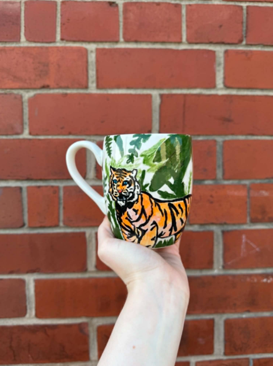 A white hand holding a ceramic mug with a hand-painted tiger and green foliage, against a red brick wall background.
