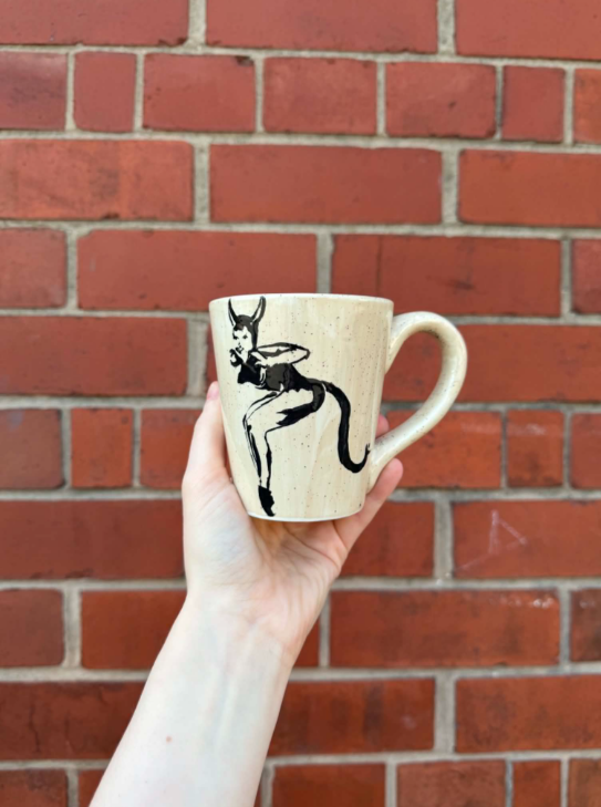 A hand holding a beige ceramic mug with a black illustration of an anthropomorphic devil or demon figure against a red brick wall background.