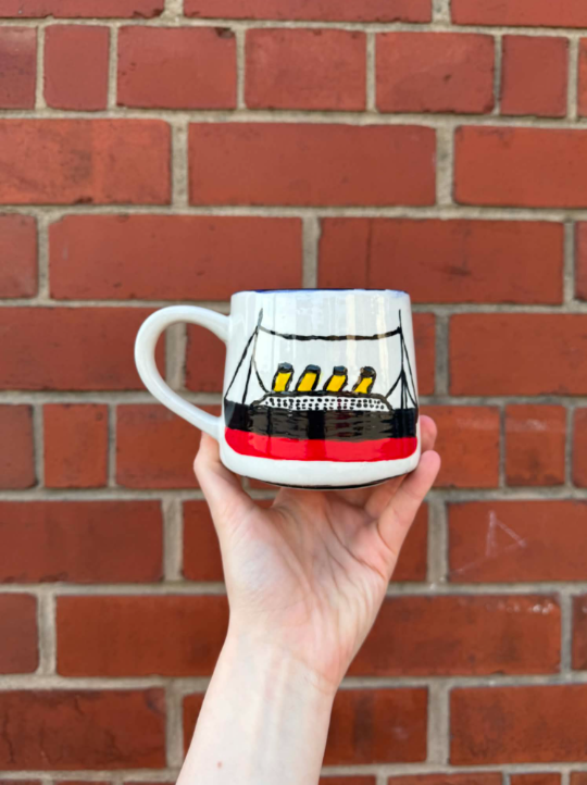 A hand holding a coffee mug painted to look like a ship, with a brick wall in the background.