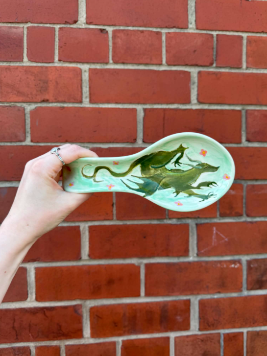 Hand holding a ceramic spoon painted with a dragon and clouds against a red brick wall background.