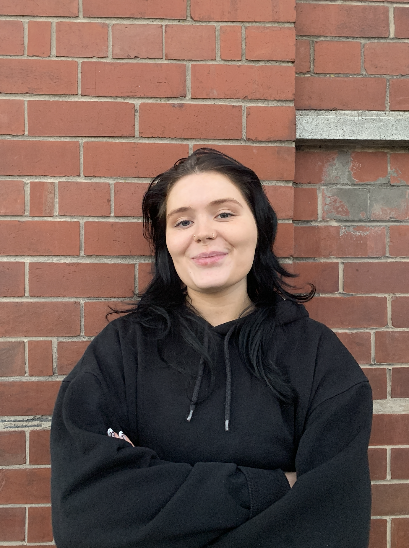 A young person with black hair and light skin, wearing a black hoodie, standing in front of a red brick wall with her arms crossed and smiling at the camera.