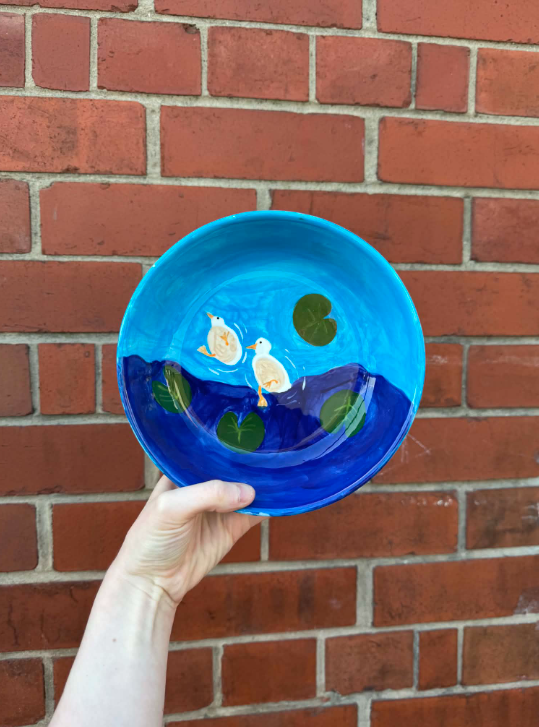 Decorative ceramic plate with painted ducks and lily pads, held against a brick wall.