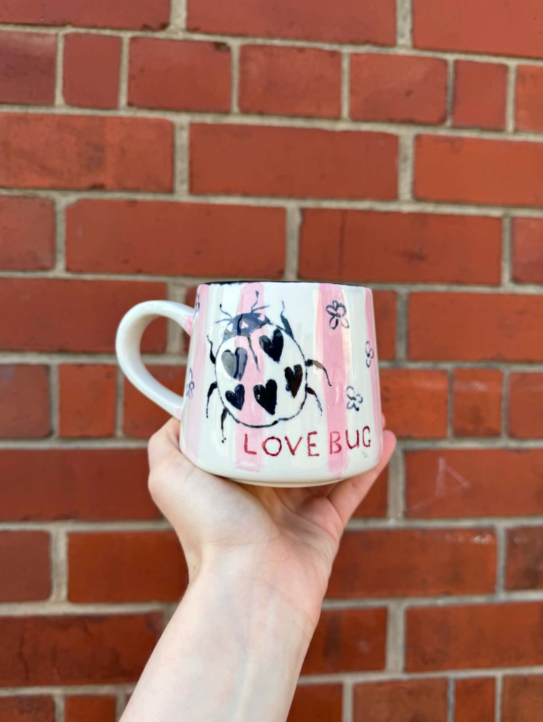 Hand holding a ceramic mug with a drawing of a bug with heart-shaped spots and the words "LOVE BUG" in red, against a red brick wall background.
