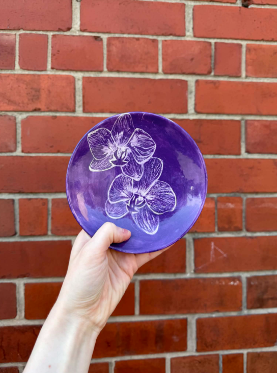 A person holding a purple plate with white orchid flower illustrations against a red brick wall background.