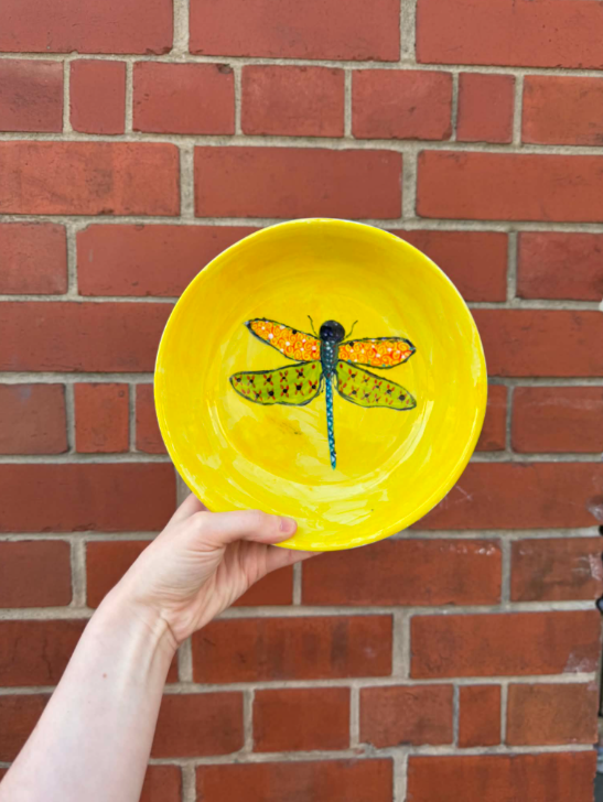 Yellow ceramic bowl with a painted dragonfly in the center, held by a person's hand in front of a brick wall.