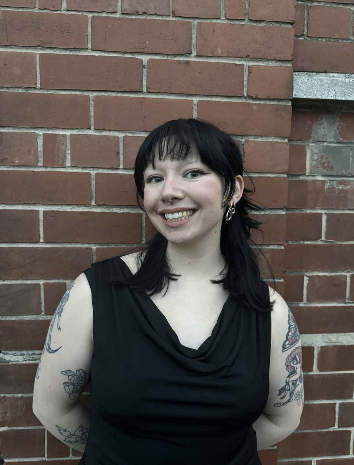Young person with black hair, tattoos on both arms, earrings, and a septum piercing, smiling in front of a brick wall.