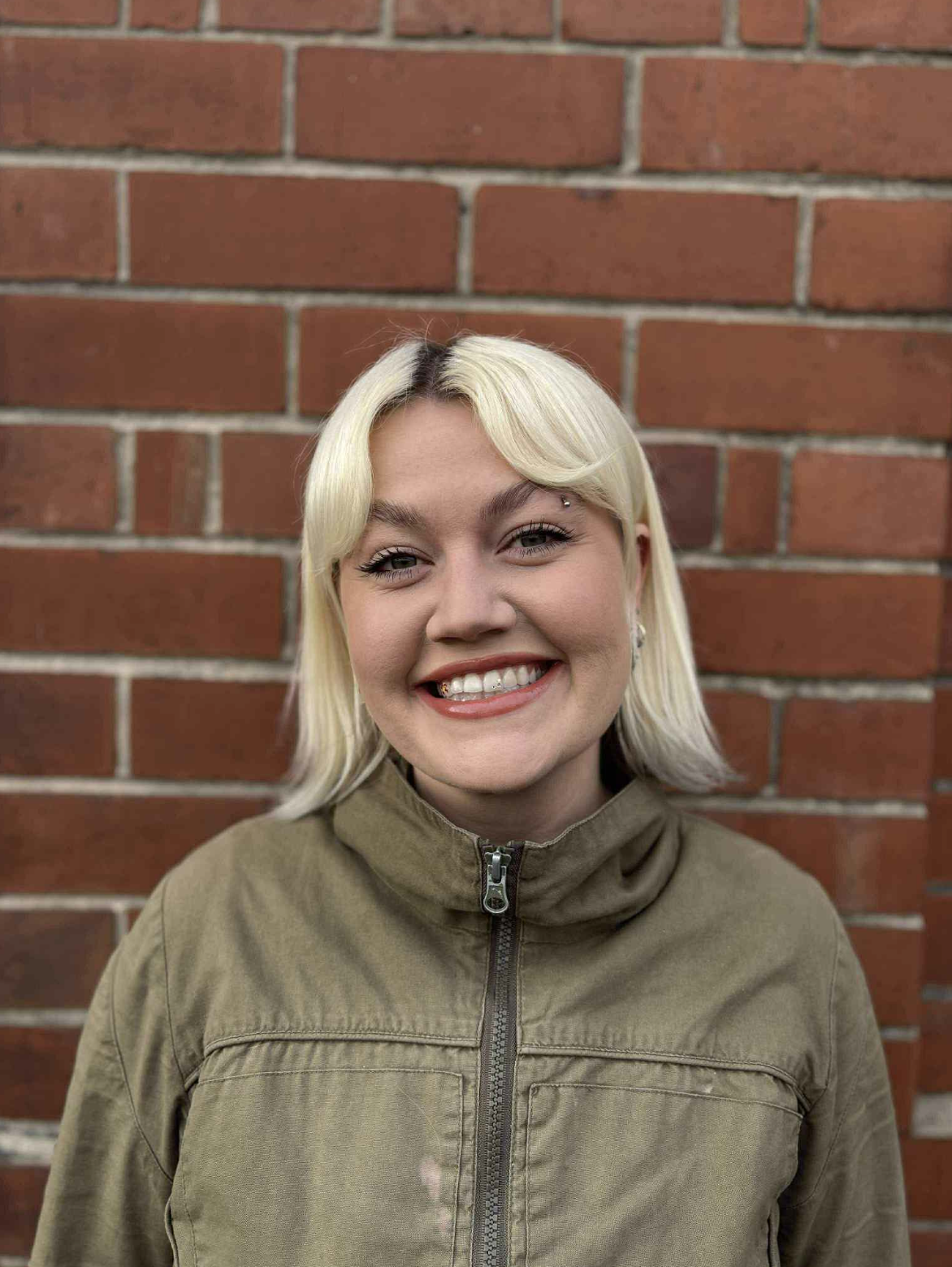 A young person with blonde hair smiling in front of a red brick wall, wearing a tan jacket with a zipper.