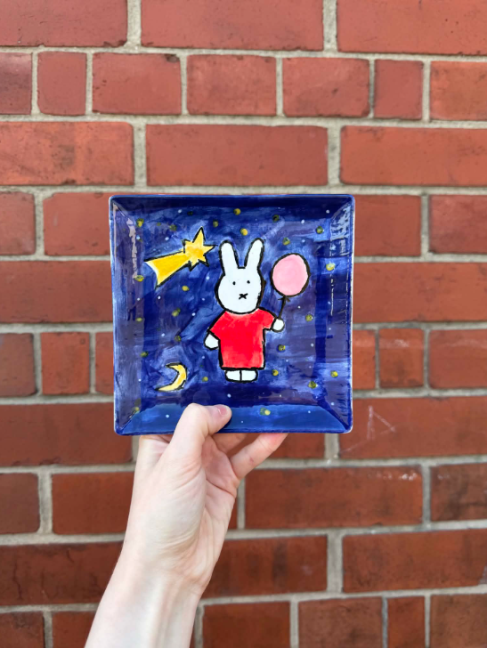 A small square ceramic dish with a hand-painted illustration of a white bunny in a red dress holding a pink balloon, against a dark blue background with stars, a shooting star, and a crescent moon, held in front of a red brick wall.