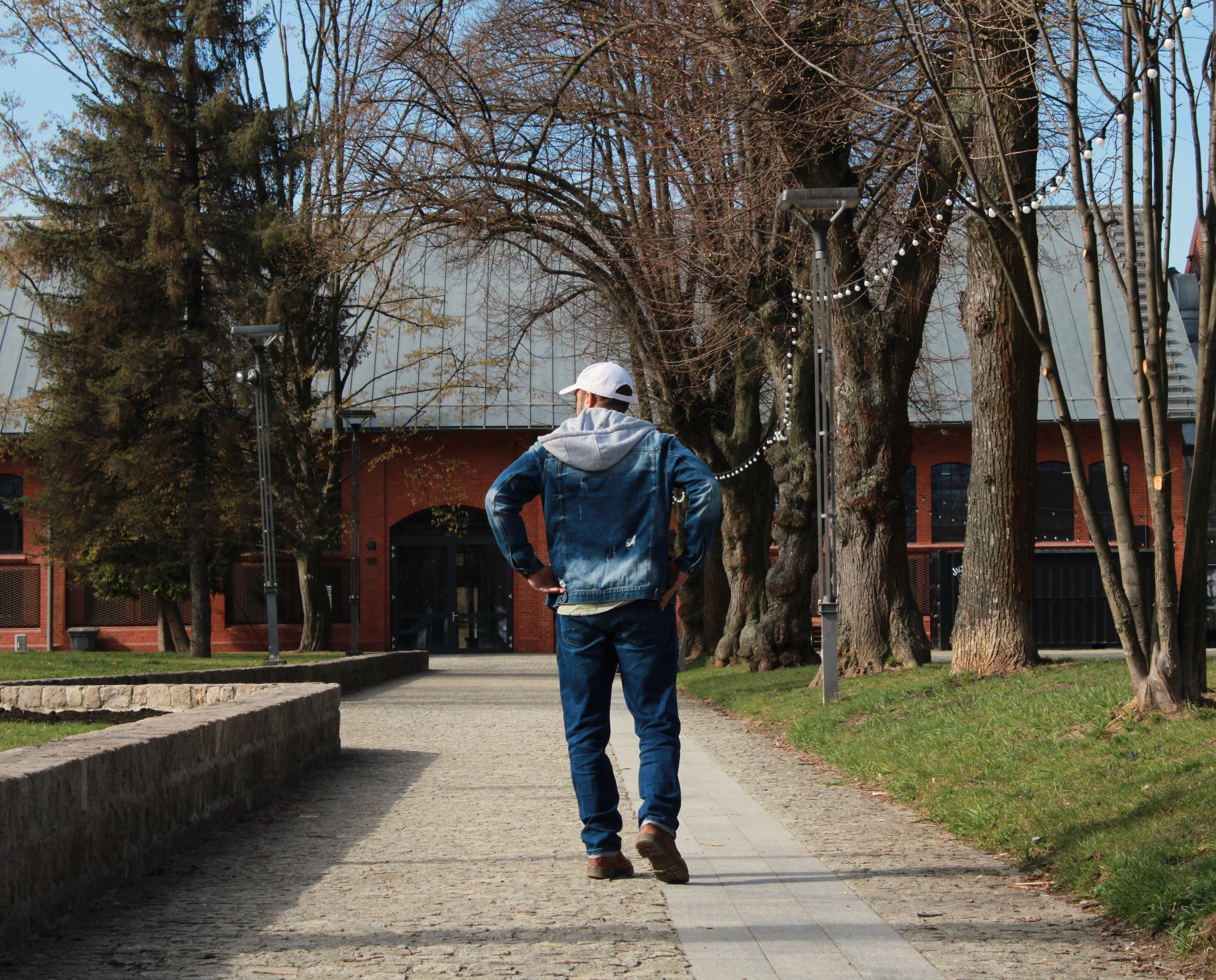 A person wearing a white cap, denim jacket, and jeans standing on a street in a park with leafless trees and string lights hanging outdoors.