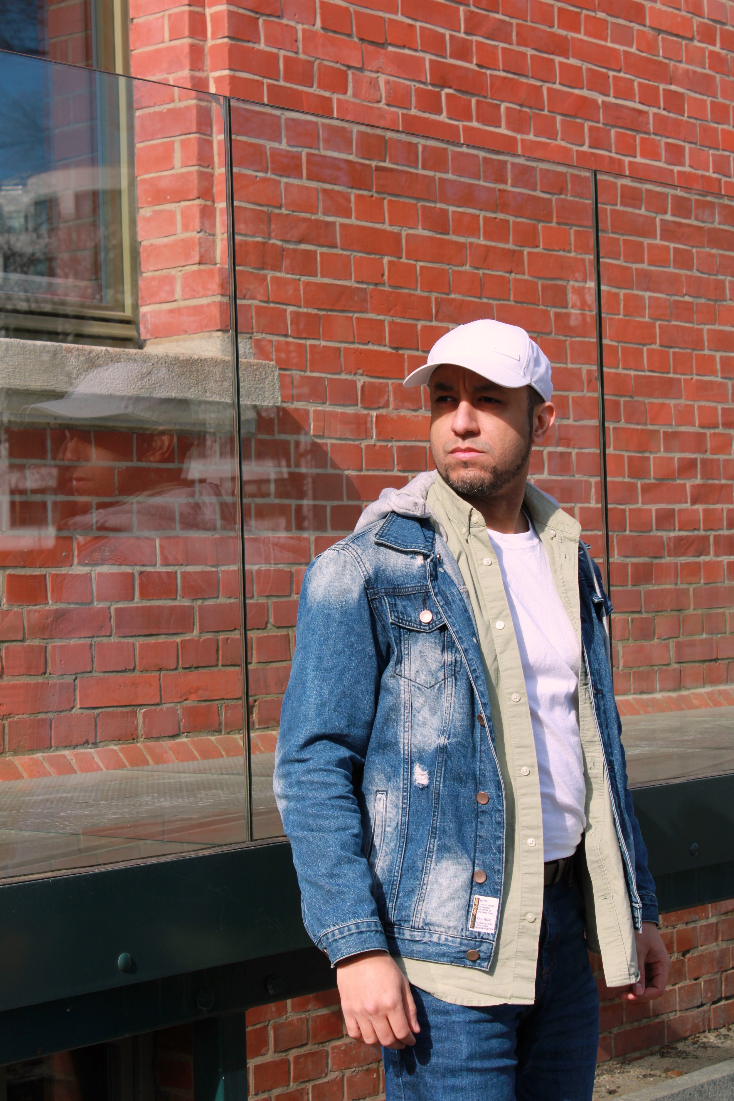 A man wearing a white baseball cap, denim jacket, and beige hoodie stands outside against a brick wall with a glass window.