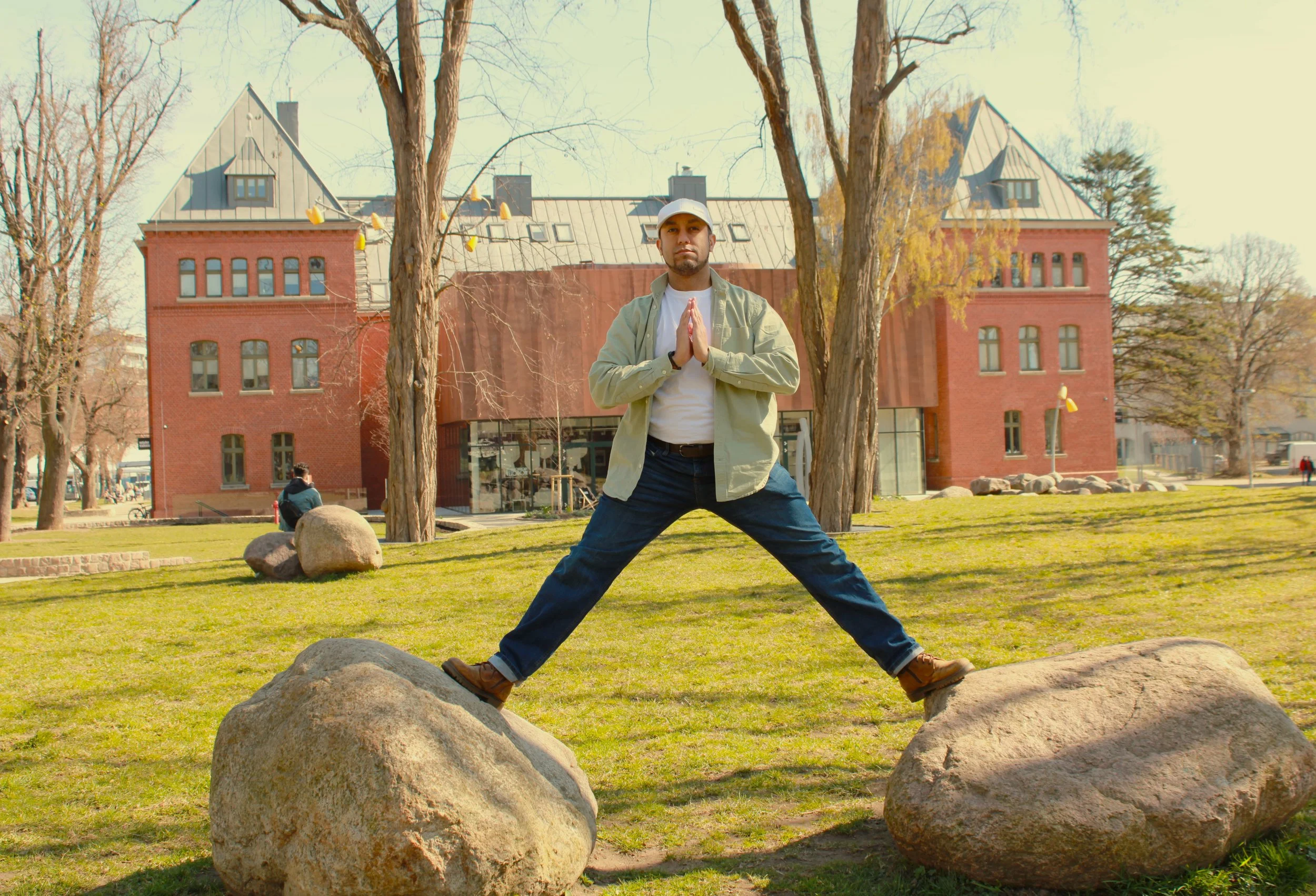 A man practicing yoga outdoors on rocks in a park with a red brick building and leafless trees in the background.