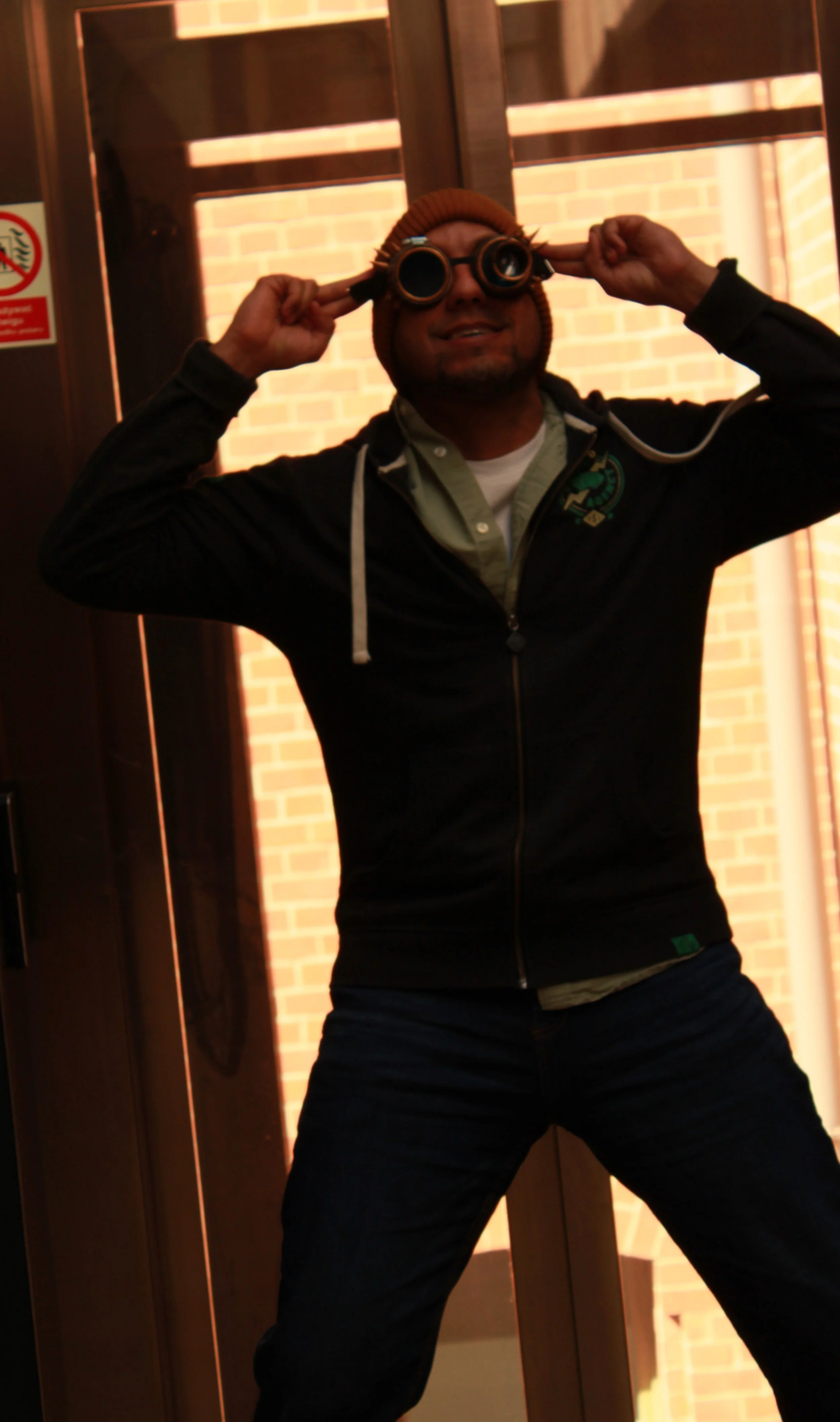 Man wearing a brown beanie and black jacket holding binoculars up to his eyes, standing indoors in front of a wooden door with a brick wall in the background.