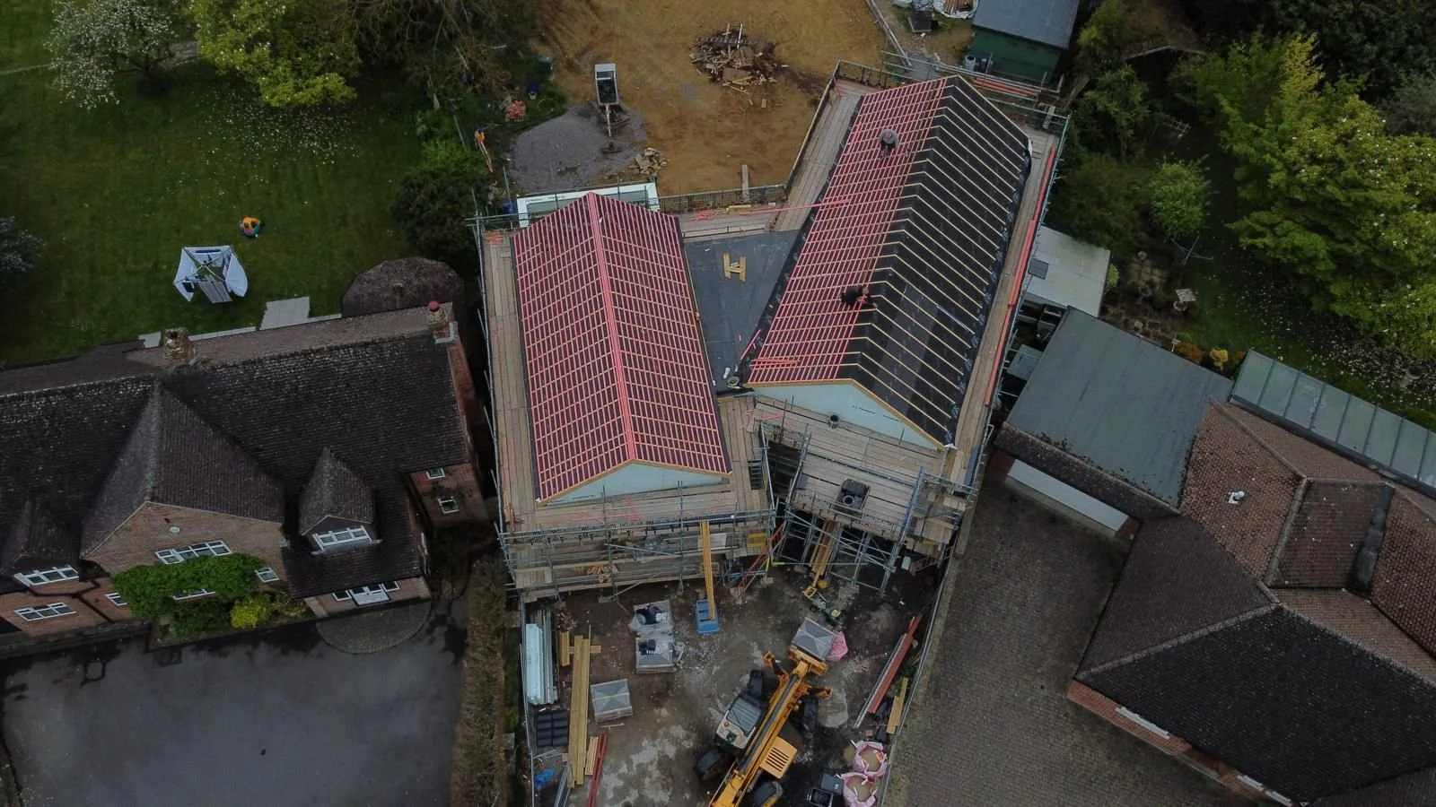 Aerial view of a building under construction with scaffolding and crews working on the roof, which has red framing. Surrounding elements include neighboring houses, a green yard with a playset, and trees.