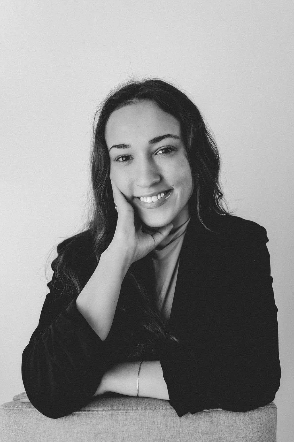 A black and white portrait of a smiling woman with long wavy hair, wearing a dark blazer, resting her chin on her hand, against a plain light background.