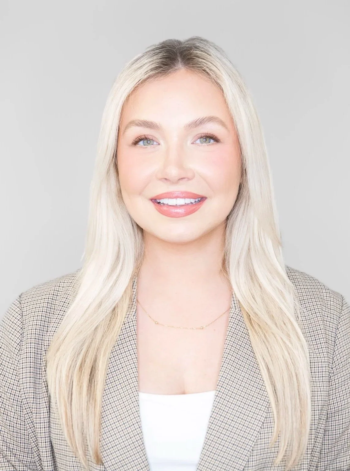 Portrait of a young woman with long blonde hair and blue eyes, smiling, wearing a beige plaid blazer and a white top, against a gray background.