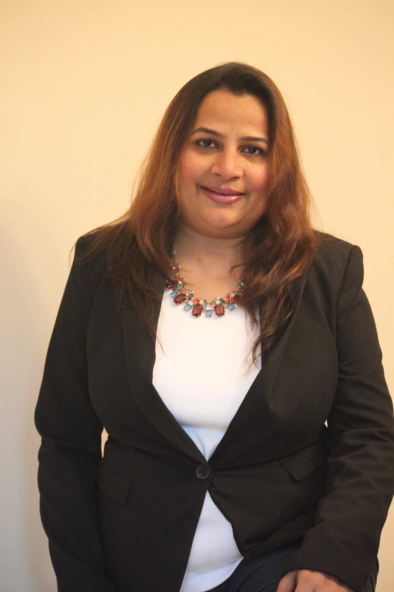 A woman with shoulder-length brown hair, wearing a black blazer, white top, and a colorful statement necklace, sitting against a plain beige wall.