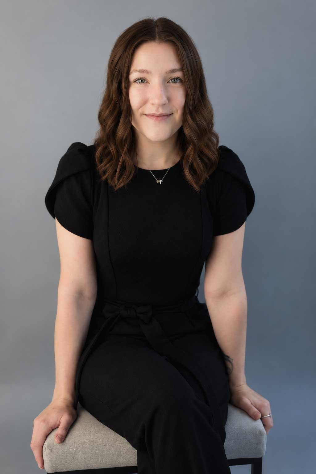 A young woman with shoulder-length wavy brown hair, wearing a black dress with puffed short sleeves, sitting on a light gray cushioned stool against a plain gray background, smiling slightly.