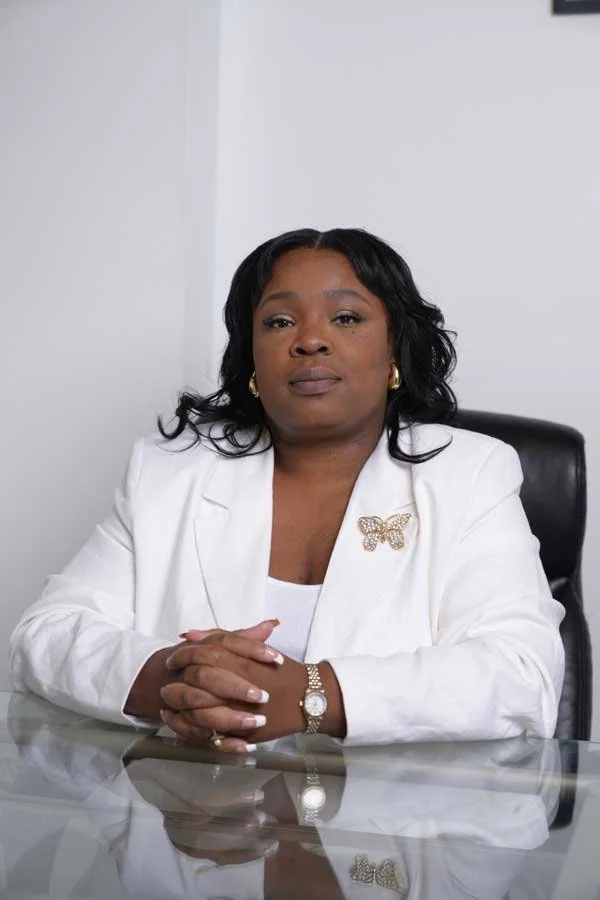 A woman sitting at a glass table in an office, wearing a white blazer with a butterfly pin, gold earrings, and a gold watch, with her hands folded on the table.