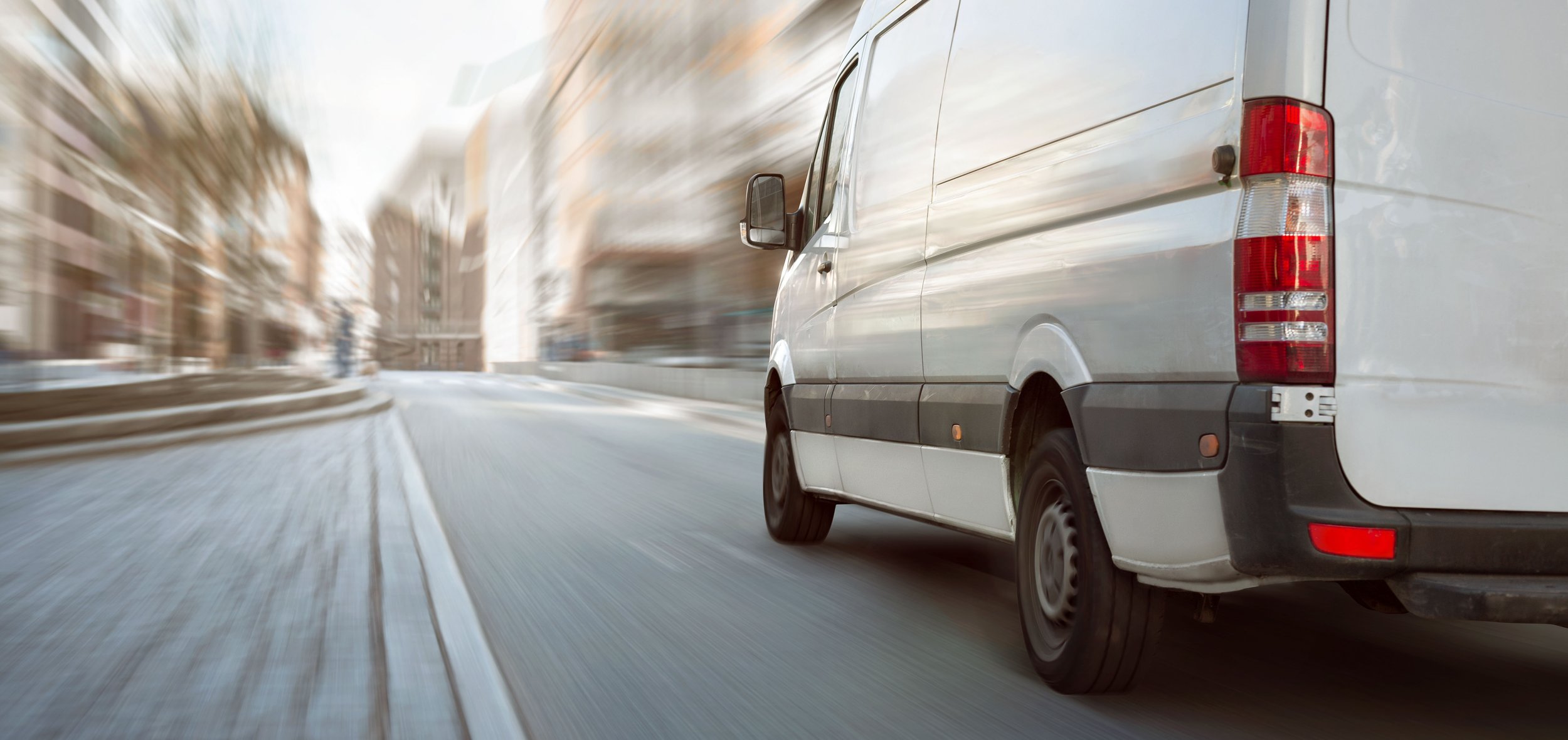 A white delivery van driving quickly through an urban street with blurred buildings and scenery around, creating a sense of motion.