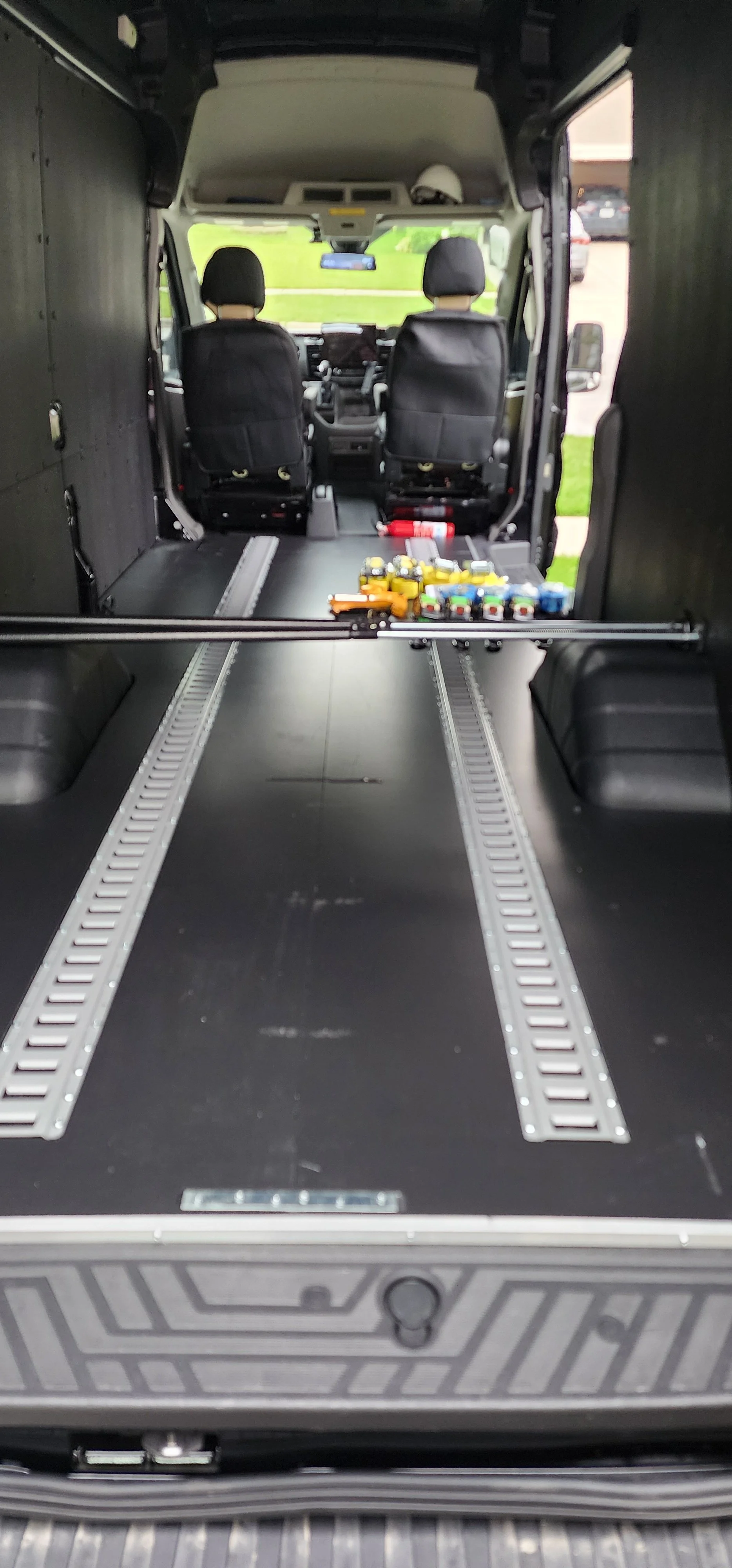 Empty black van interior with tracks on the floor, with cargo tie-down straps lined up by the side door of the cargo area, viewed from the open rear door.