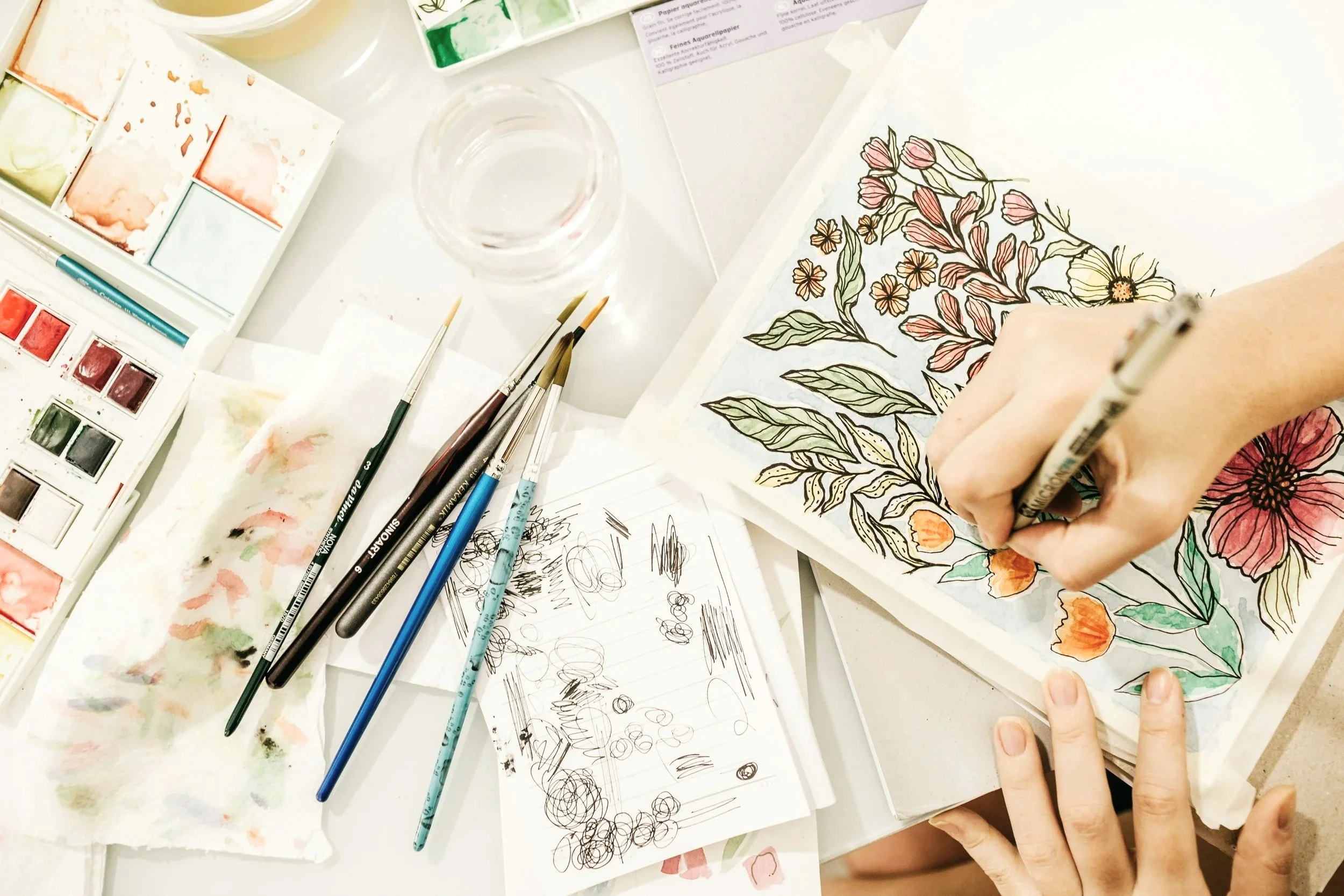 two hands at a desk, painting flowers onto a sketchpad with painting supplies on the desk