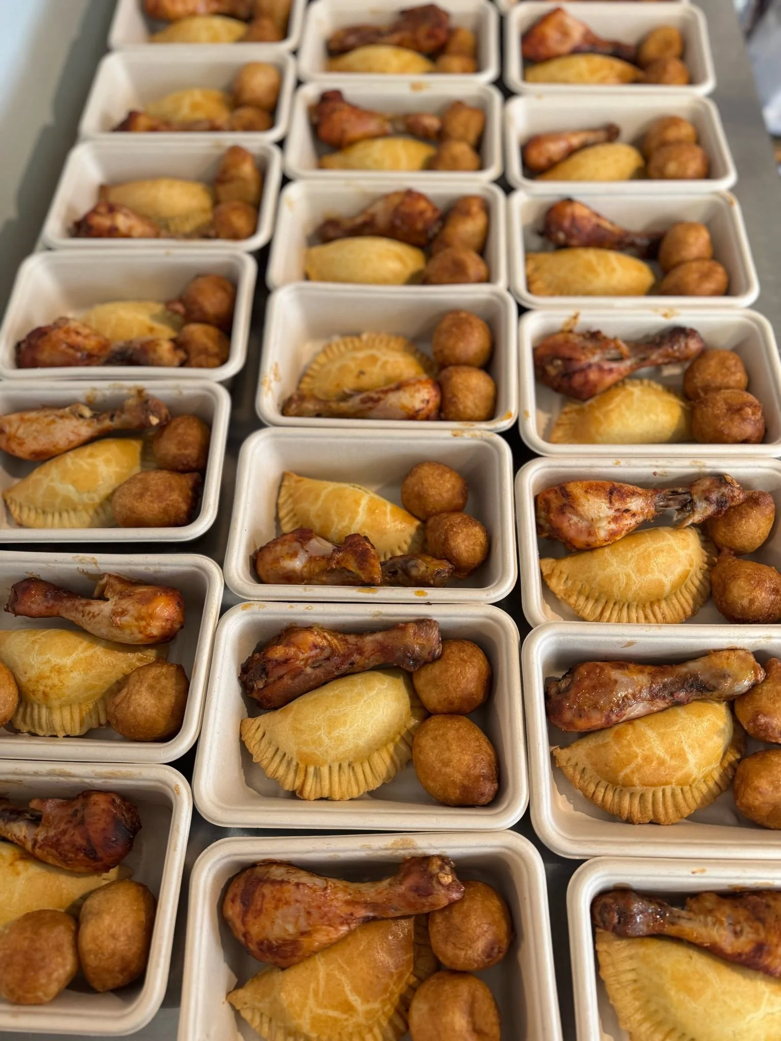 Multiple white takeaway containers filled with fried chicken drumsticks, meat pies, and puff puff arranged on a table.