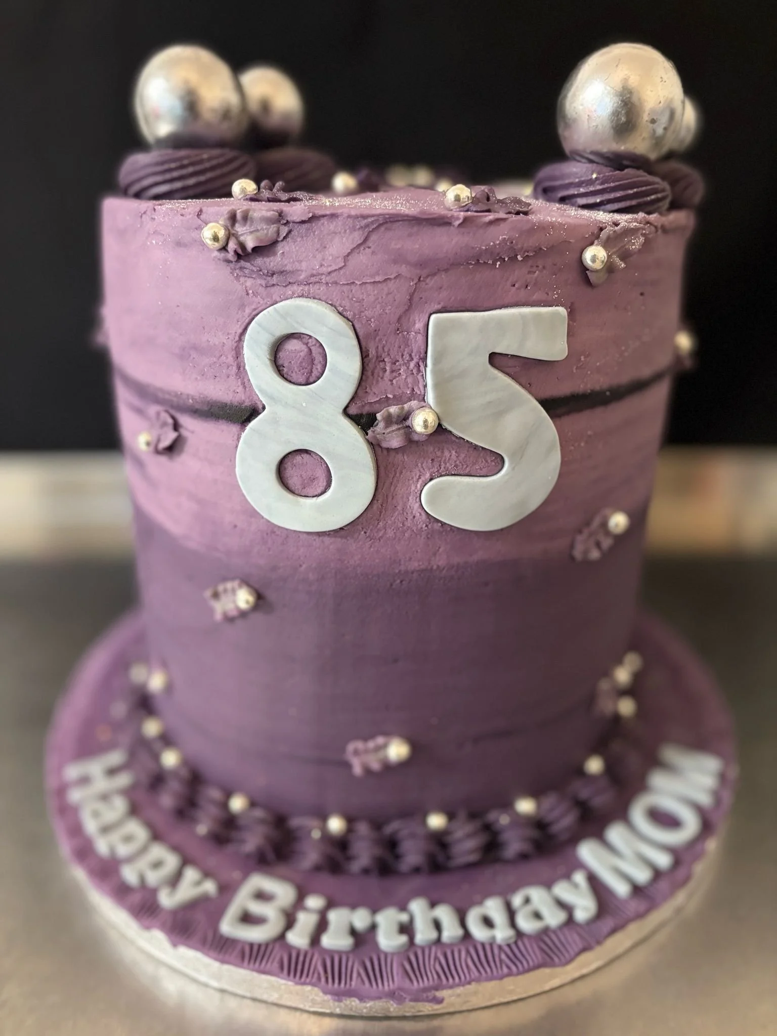 A purple birthday cake decorated with silver and purple balls, purple frosting, and white '85' numbers on top.