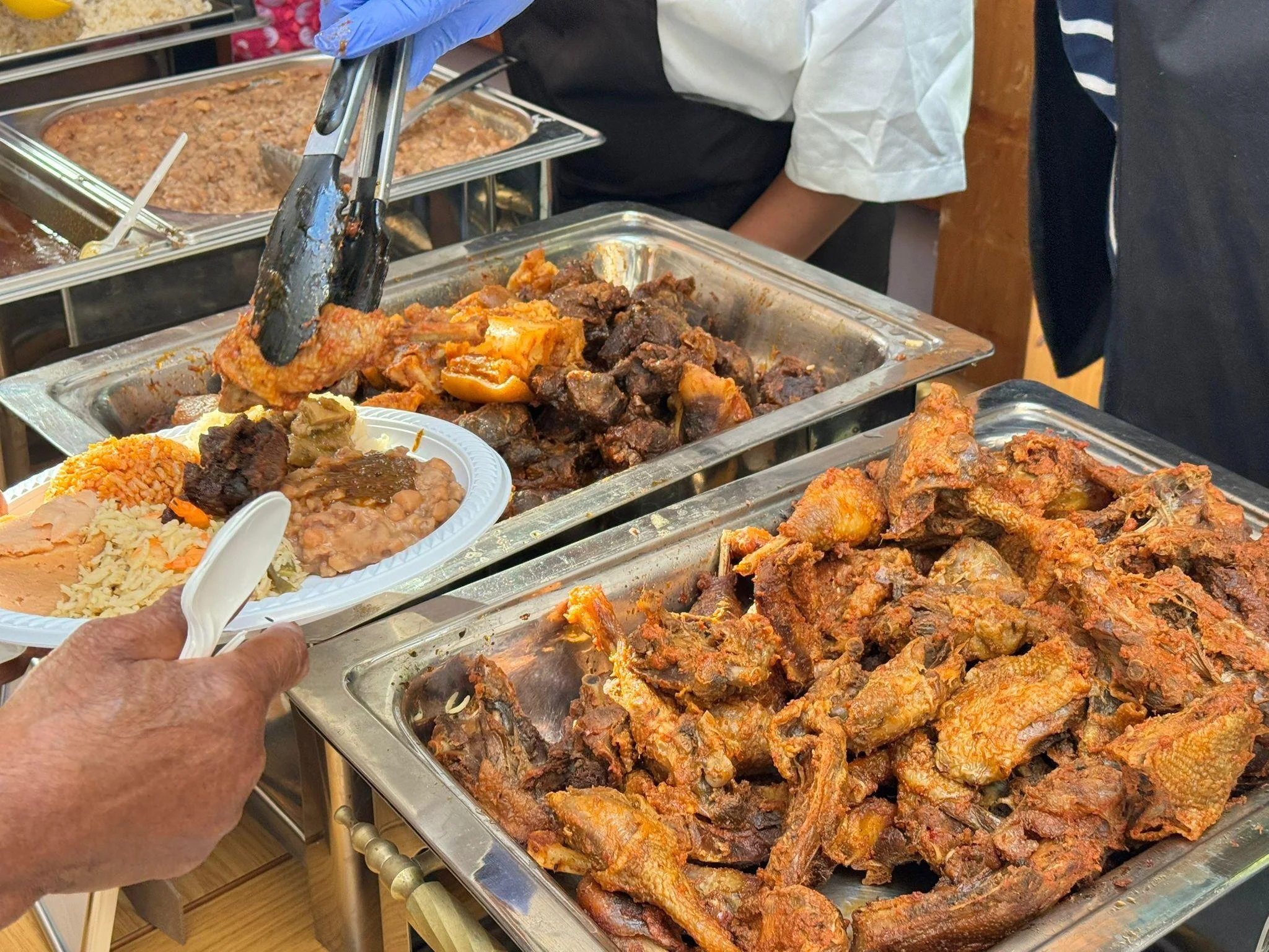 Steel buffet trays filled with various cooked meats and side dishes, including ribs and fried fish, at a food service setting, with a person using tongs to serve food.