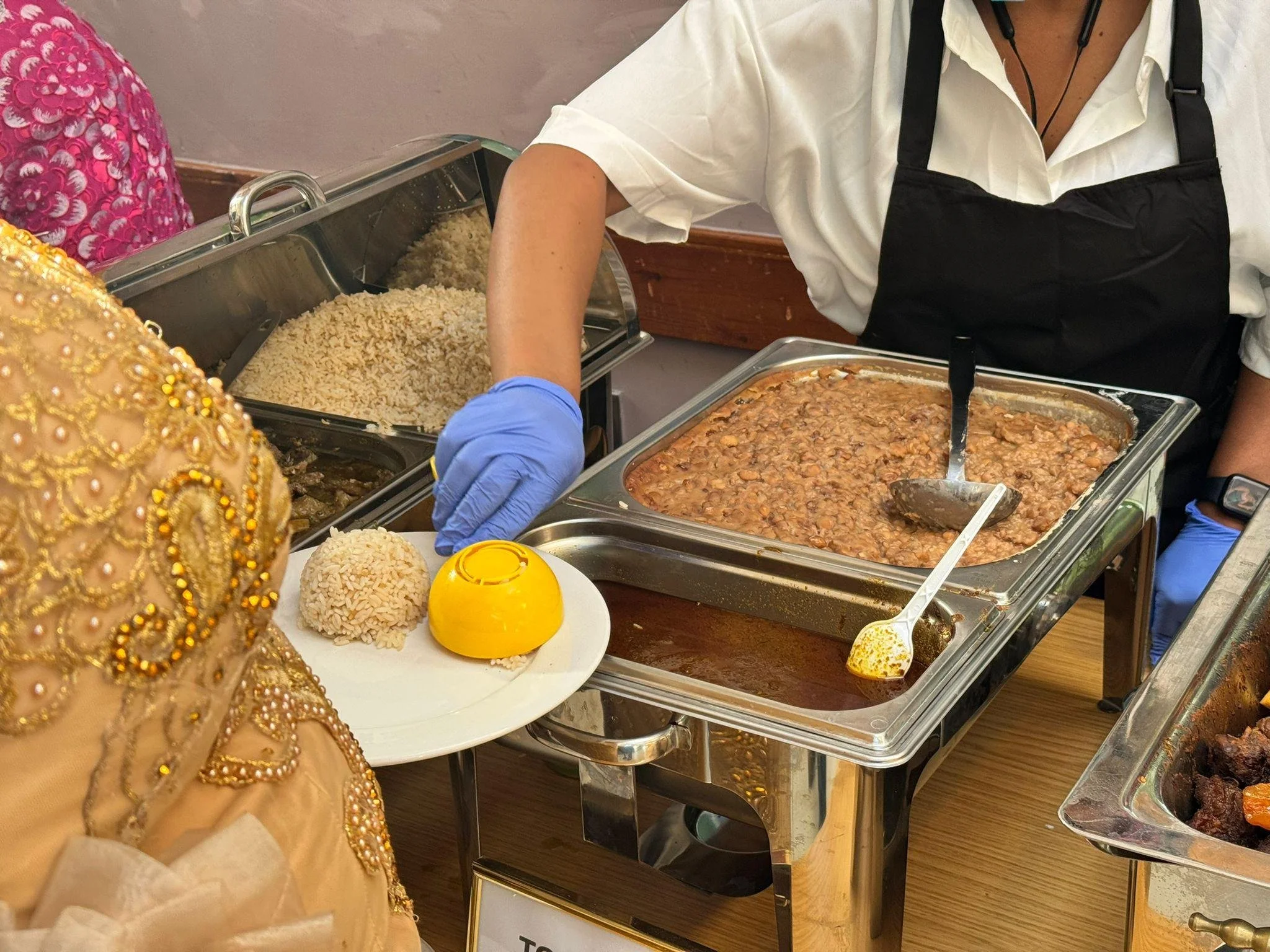 A buffet table with serving trays of rice and beans, and a guest in a black apron serving food. A person in colorful, ornate clothing is partially visible in the foreground.