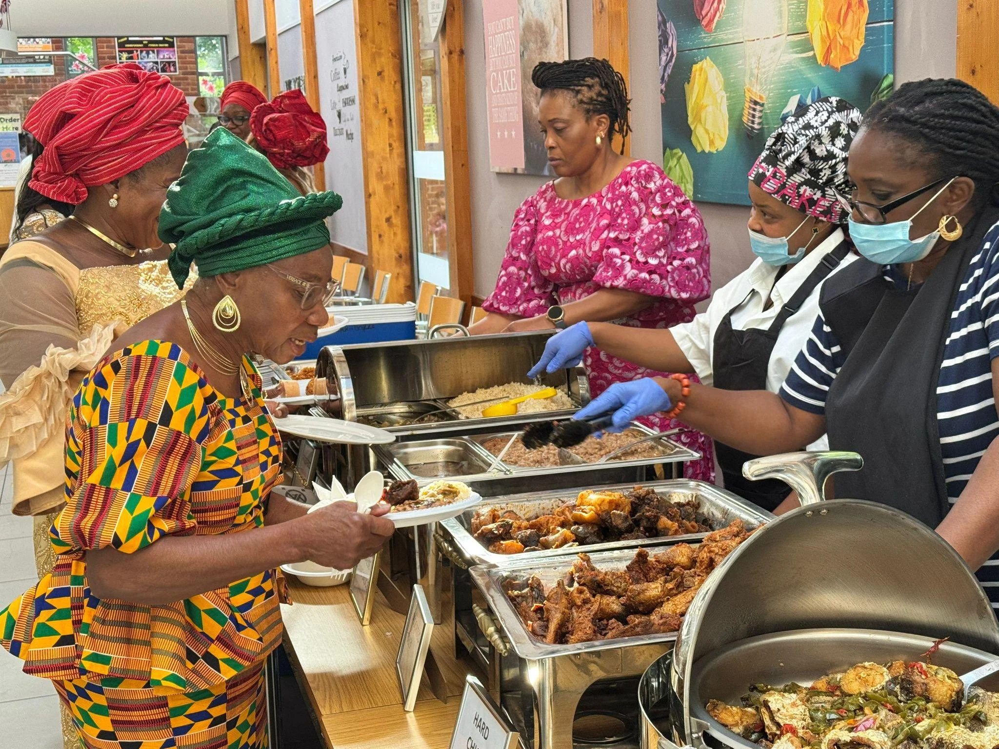 Women and a man serving themselves food at a buffet with various dishes, including fried chicken. Women are wearing colorful African attire and headwraps, while the man is wearing a striped shirt and medical mask.