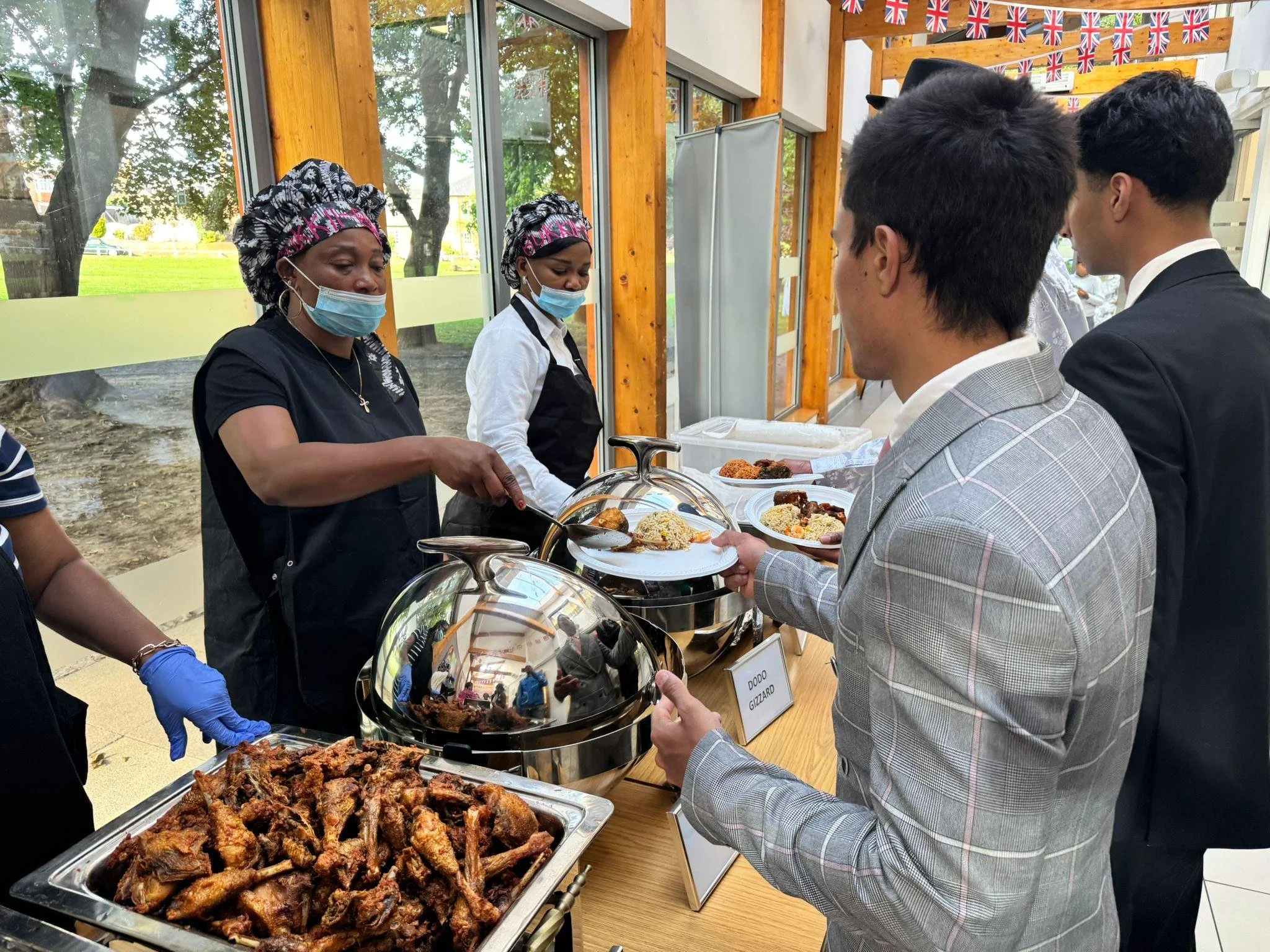 People selecting food from a buffet table at a cultural event, with British flags hanging in the background.