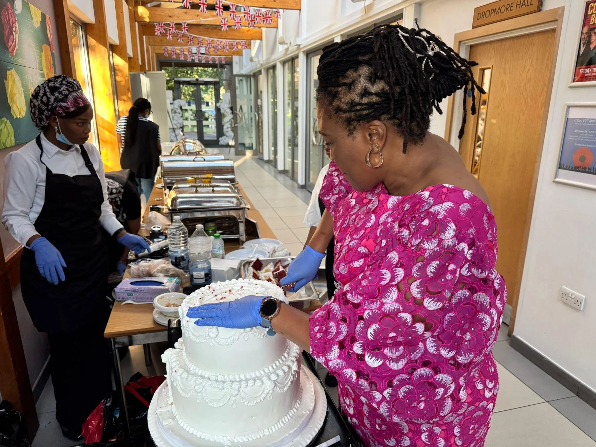 A woman in a pink patterned dress with black dreadlocks decorated with pearls, wearing a watch and earrings, appears to be serving or preparing food at a buffet table. Other women wearing masks and aprons are also at the table, which has trays of foo